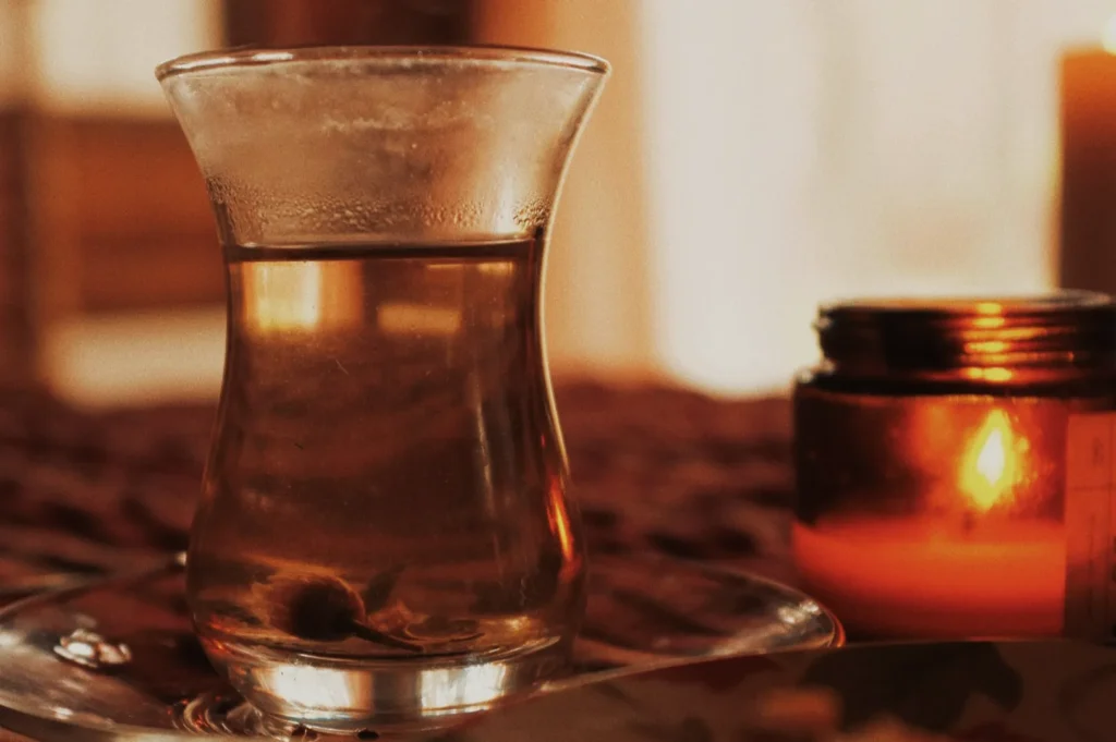 A steaming glass of tea sits on a saucer beside a lit, amber-glowing candle, creating a warm and cozy ambiance on a textured surface.