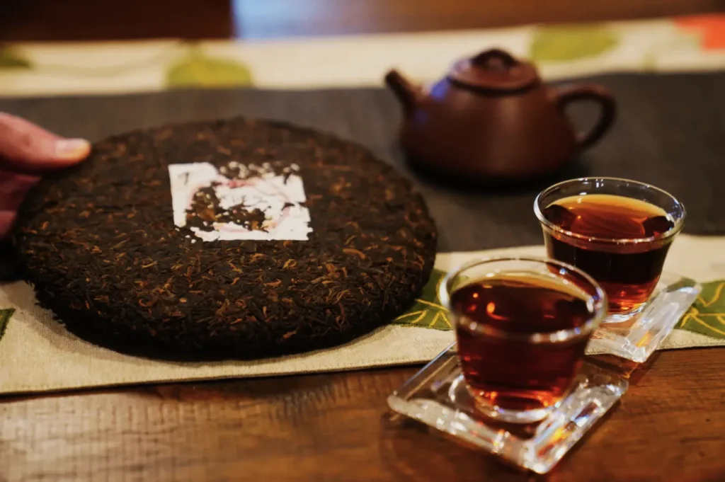 A hand holds a round compressed tea cake near a wooden table with a brown teapot and two glass cups of deep amber tea, creating a warm, inviting scene.