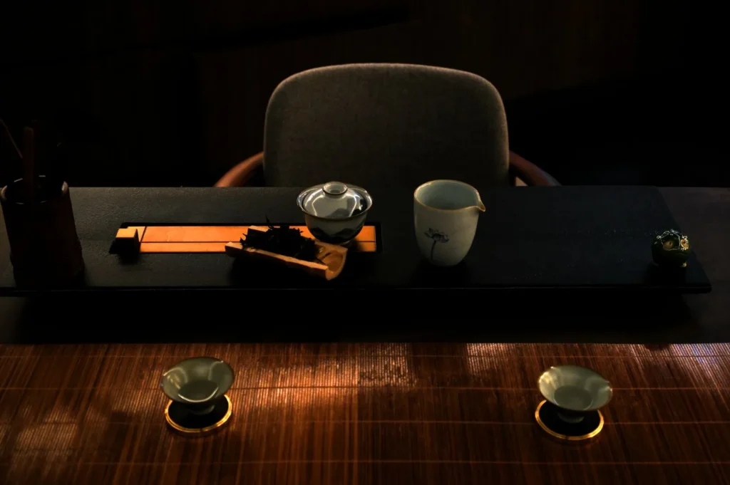 Dimly lit tea setup on a wooden table with teacups, a teapot, and utensils. A gray chair is in the background, creating a serene atmosphere.