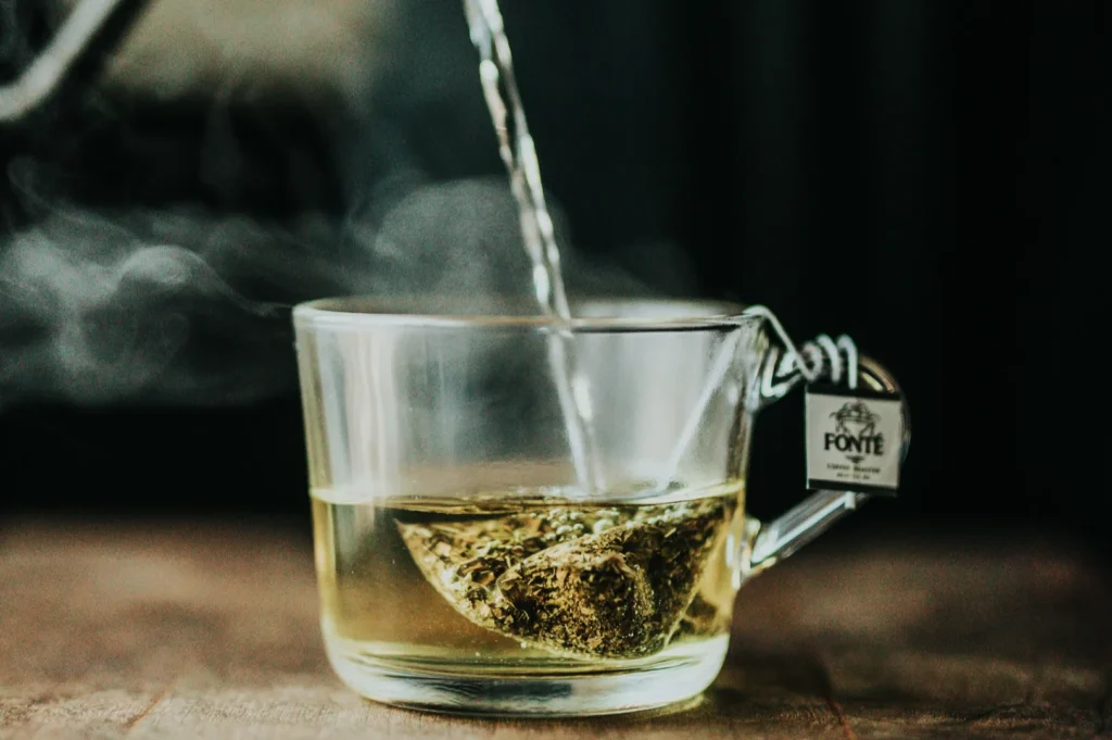 Clear glass mug with a tea bag inside, being filled with hot water. Steam is rising, and a tag hangs from the handle, creating a warm, calming mood.