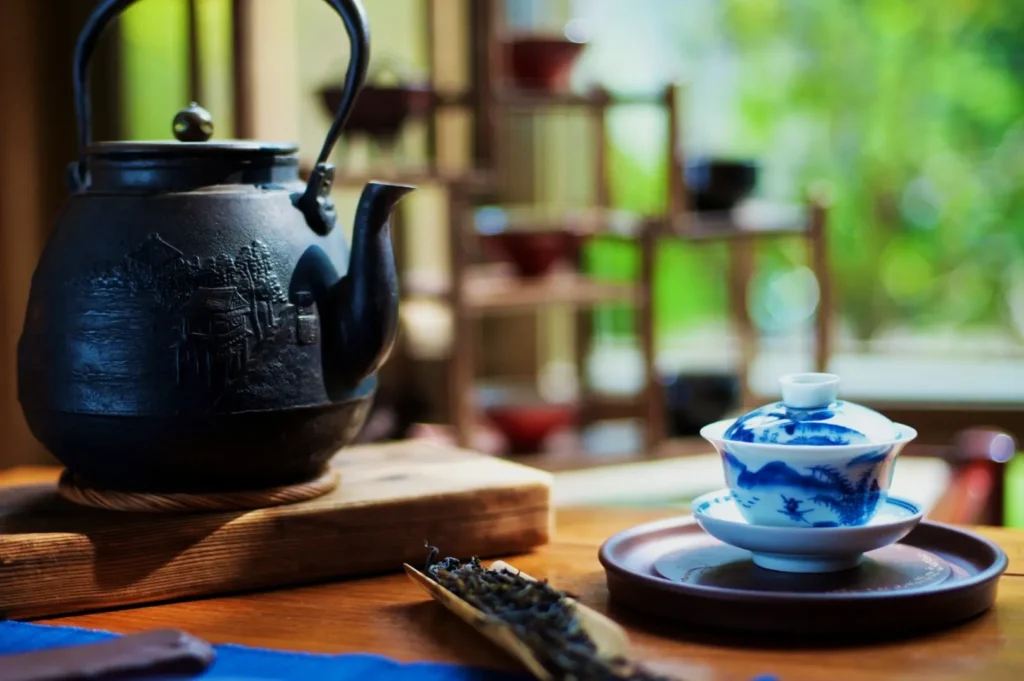 A black cast iron teapot sits on a wooden board beside a blue and white porcelain tea set. Loose tea leaves are visible in a scoop. The background has soft, natural light filtering through a window, creating a serene, inviting atmosphere.