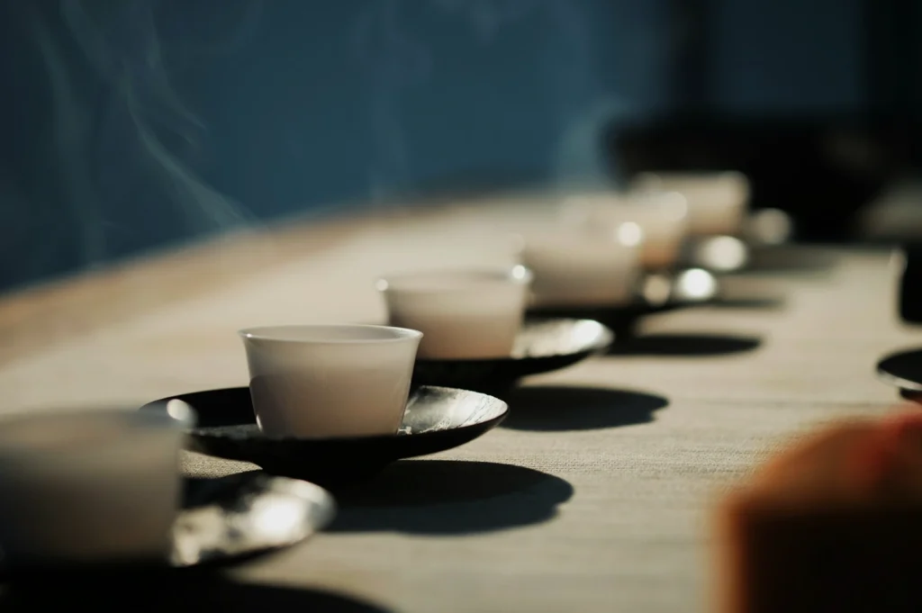 A row of white teacups on black saucers sits on a sunlit table, casting soft shadows. Steam rises gently, evoking a calm and peaceful atmosphere.