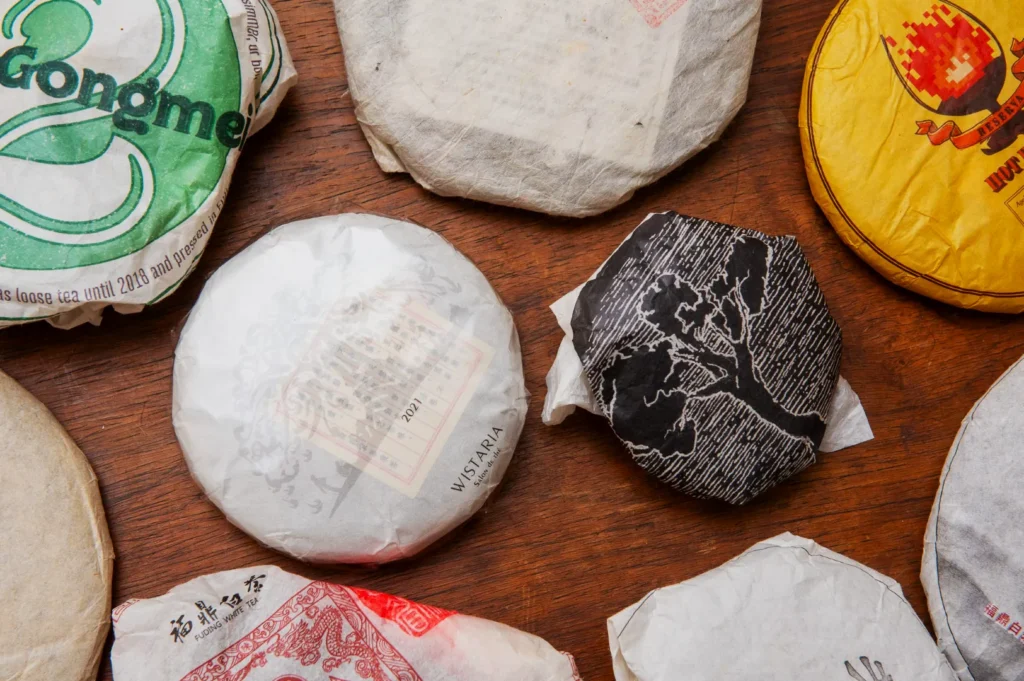 Assorted wrapped tea cakes on a wooden table. Each package has unique designs, logos, and text, representing different tea brands and origins.
