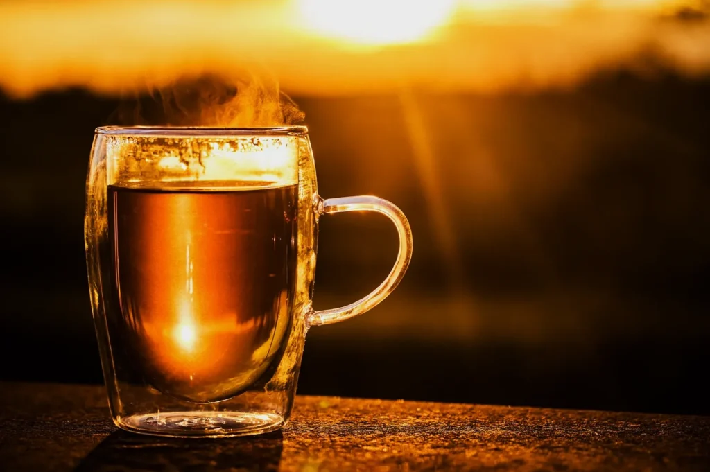 A steaming glass mug of tea sits on a surface, illuminated by a warm, golden sunset in the background, creating a cozy and serene atmosphere.