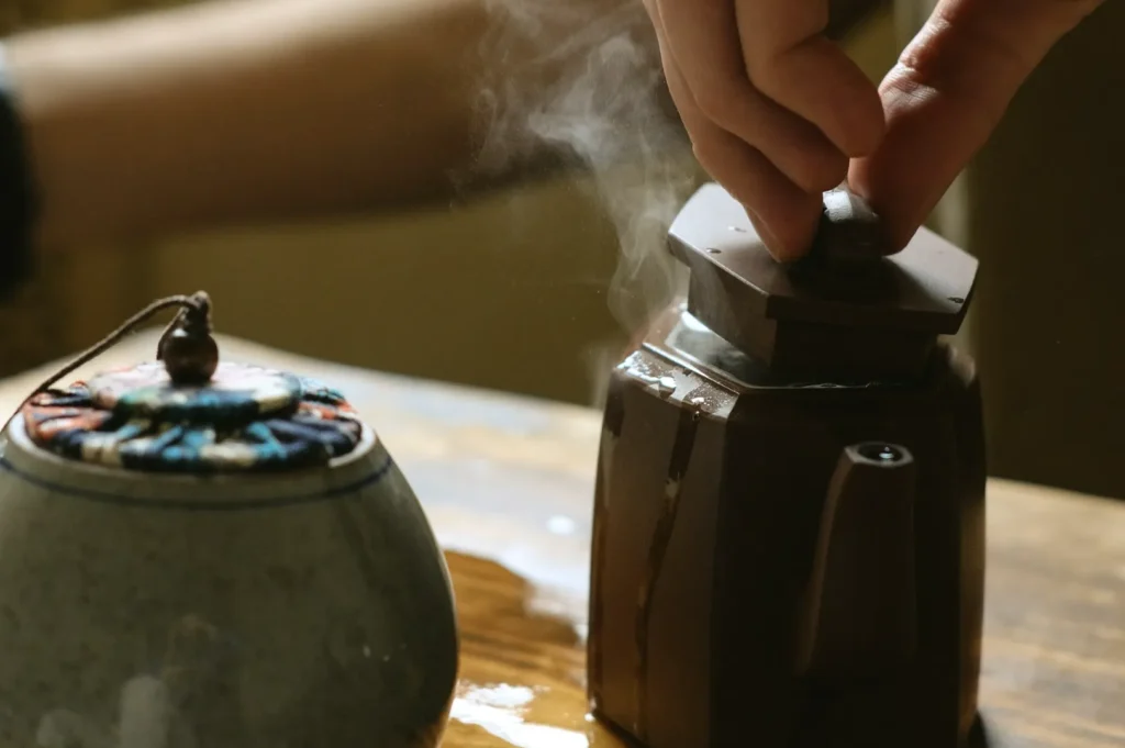A hand lifts the lid of a steaming, dark brown teapot on a wooden table. A ceramic jar with a colorful knitted cover is beside it, creating a cozy ambiance.