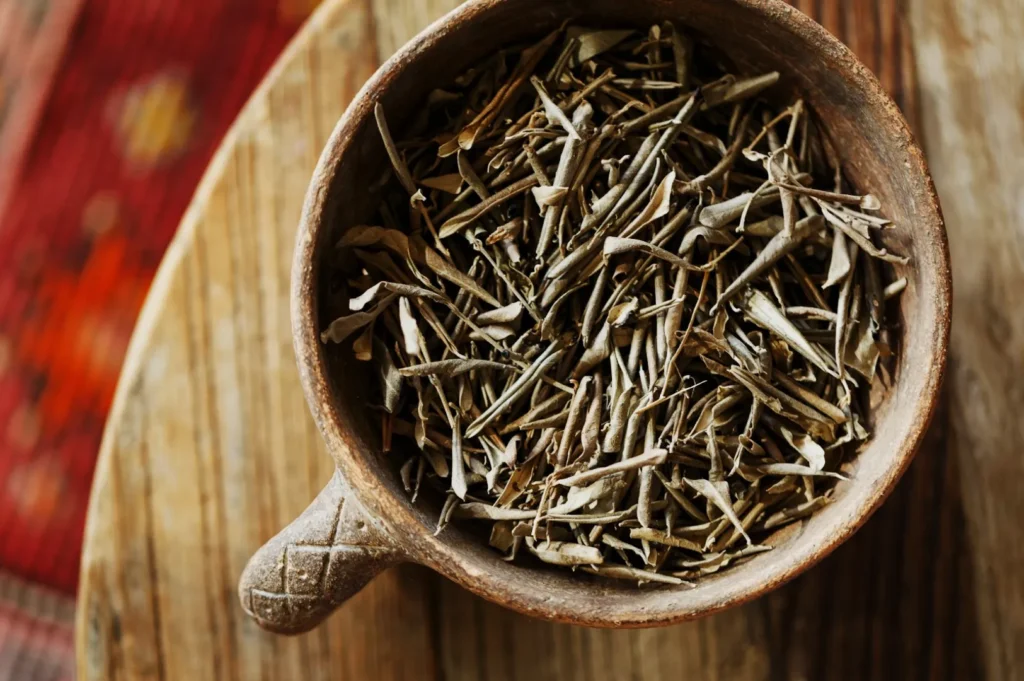 A wooden bowl filled with dried herbal sticks sits on a rustic wooden surface. The background shows a blurred red and orange patterned carpet.