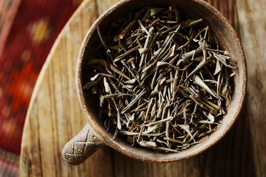 A wooden bowl filled with dried tea leaves rests on a rustic wooden surface. The background features a blurred red-patterned carpet, creating a cozy ambiance.