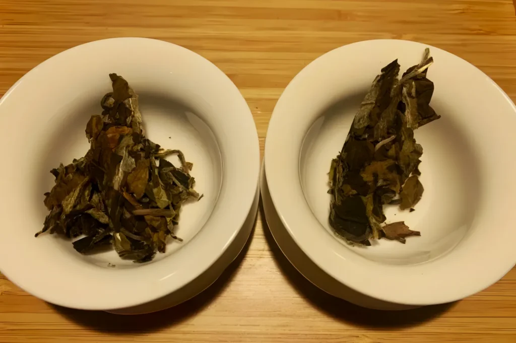Two white bowls on a wooden surface, each containing a different pile of dried leaves, showcasing variations in color and texture, suggesting a focus on tea.