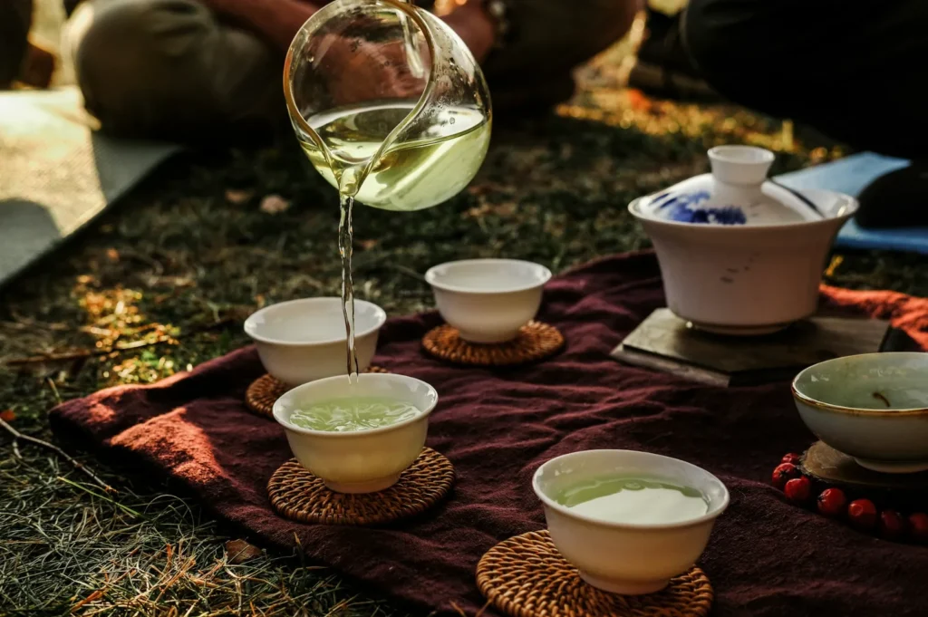 A hand pours green tea from a glass pot into white cups on woven coasters, set on a maroon cloth outdoors. Warm sunlight creates a peaceful atmosphere.