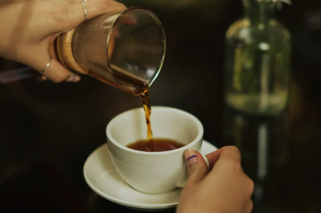 A hand pours coffee from a glass carafe into a white cup on a saucer, with a blurred glass bottle in the background, creating a cozy atmosphere.