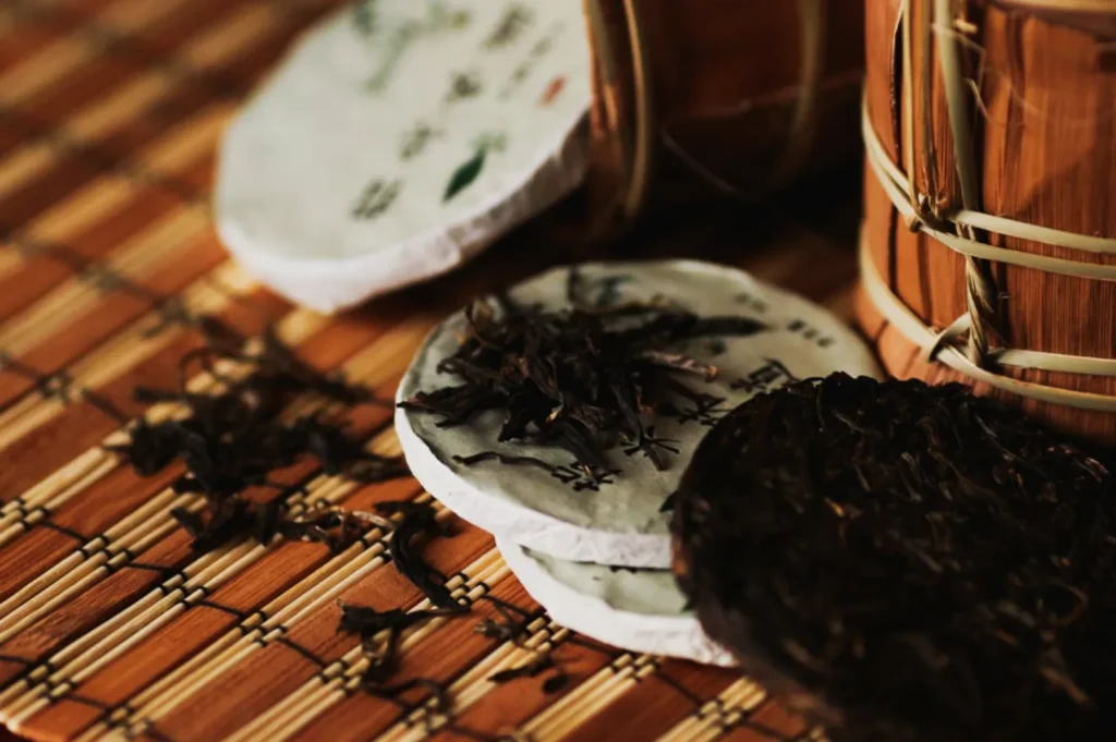 Close-up of pressed tea cakes and loose tea leaves on a woven bamboo mat. The scene conveys an earthy, rustic feel, emphasizing traditional tea culture.