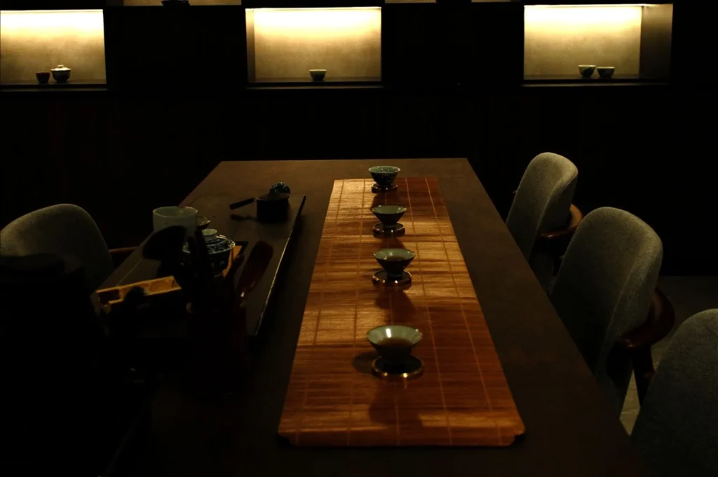Dimly lit tea room with a dark wooden table set for tea. Five cups on a bamboo mat are aligned, evoking a serene and contemplative atmosphere.