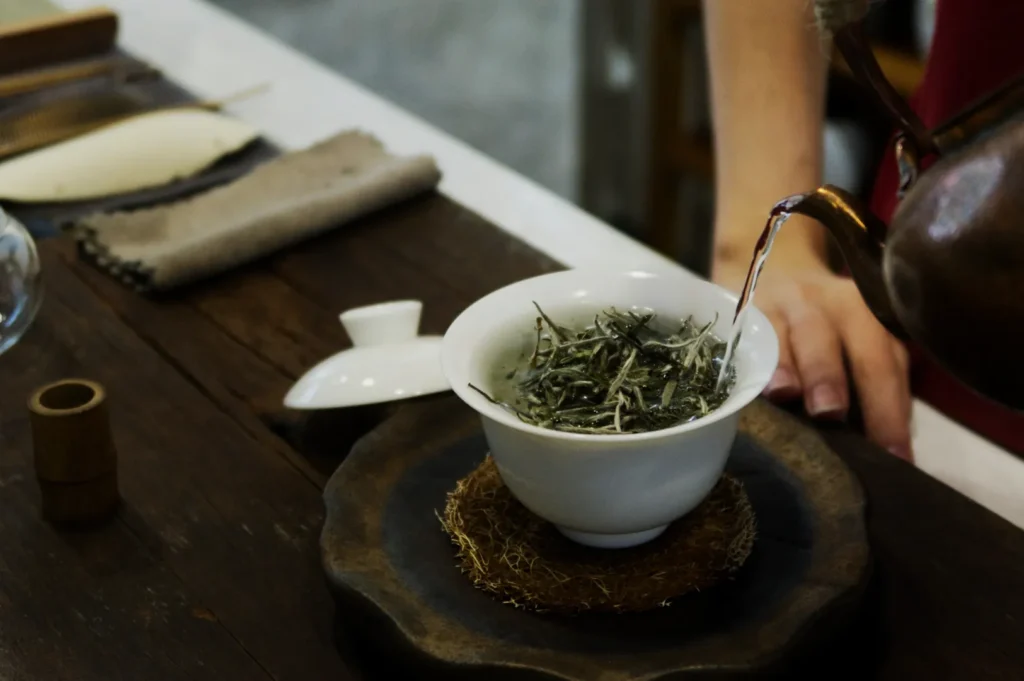 A hand is pouring water from a rustic teapot into a white bowl filled with loose tea leaves. The wooden table has a serene, inviting atmosphere.