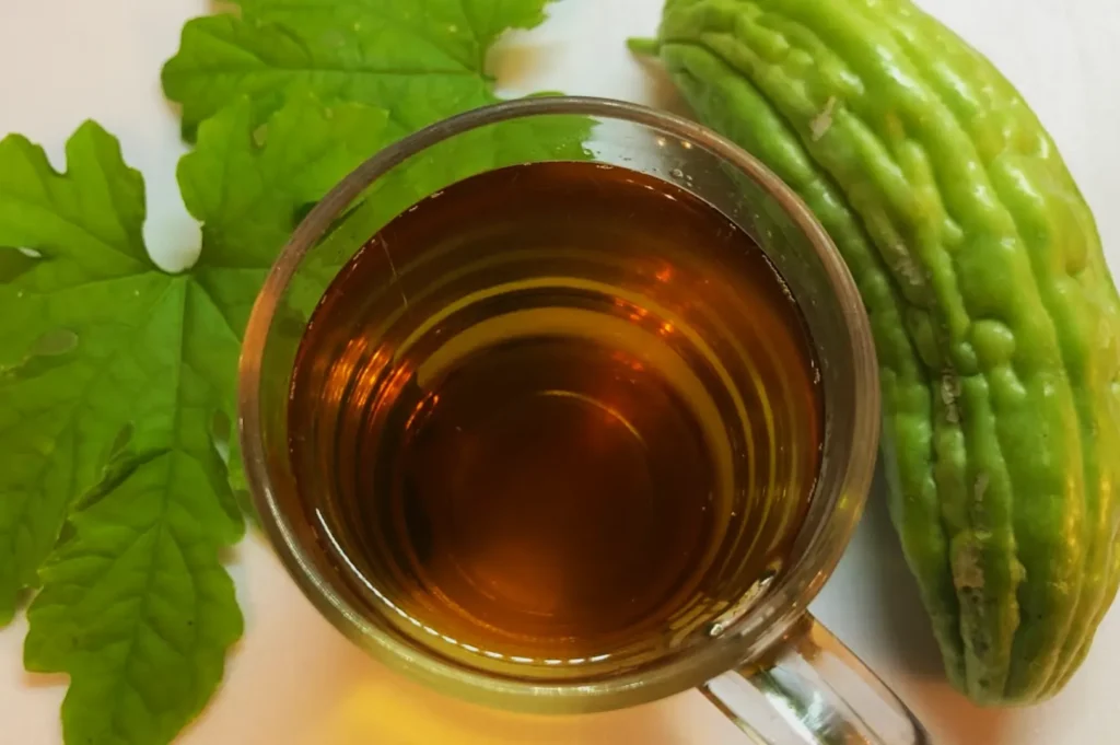 A glass of golden bitter melon tea is placed on a table, accompanied by a fresh bitter melon and bright green leaves, creating a natural, refreshing feel.