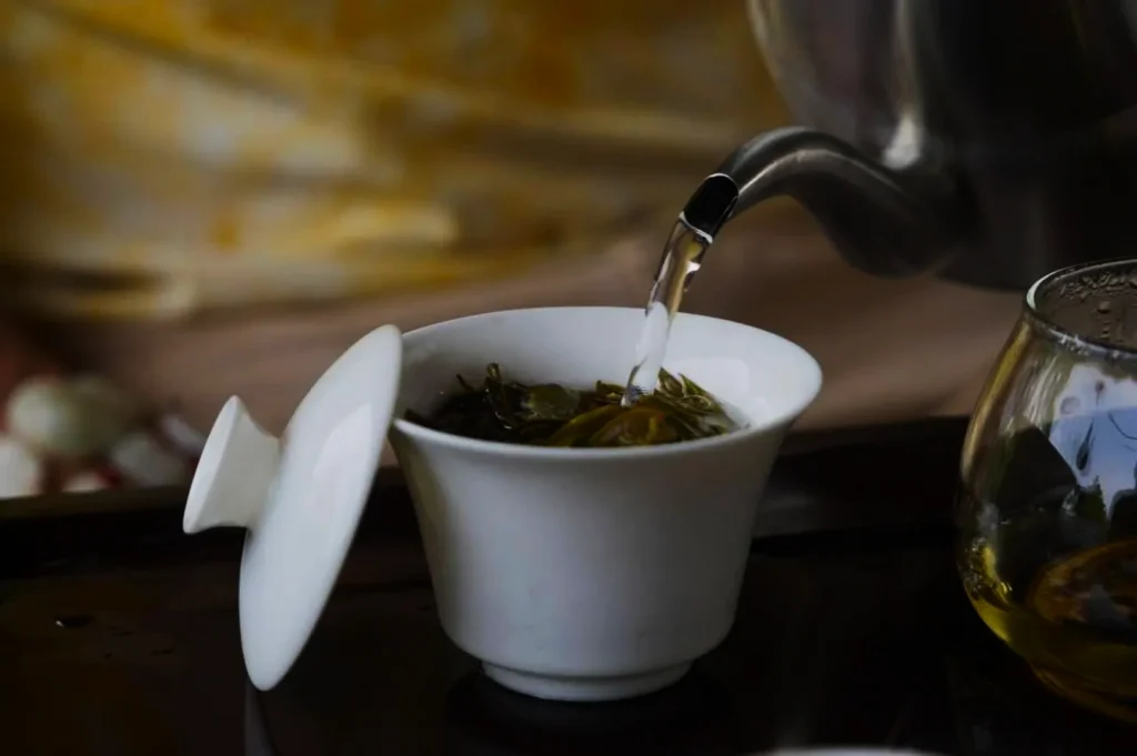 Tea being poured from a kettle into a white cup with green tea leaves, set on a tray. Nearby, a glass cup partly filled with tea. Calm and cozy atmosphere.