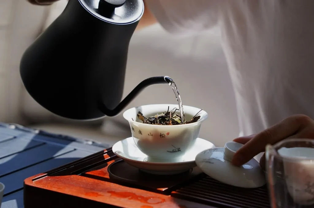 A person pours hot water from a black kettle over tea leaves in a white cup, set on a wooden tray, evoking a calm, focused atmosphere.