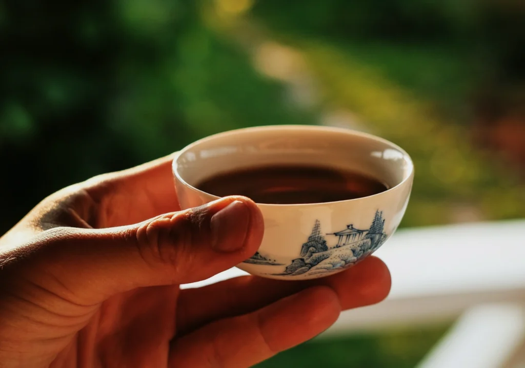 A hand holding a small porcelain teacup with blue patterns, filled with tea, against a blurred green outdoor background, conveying tranquility.