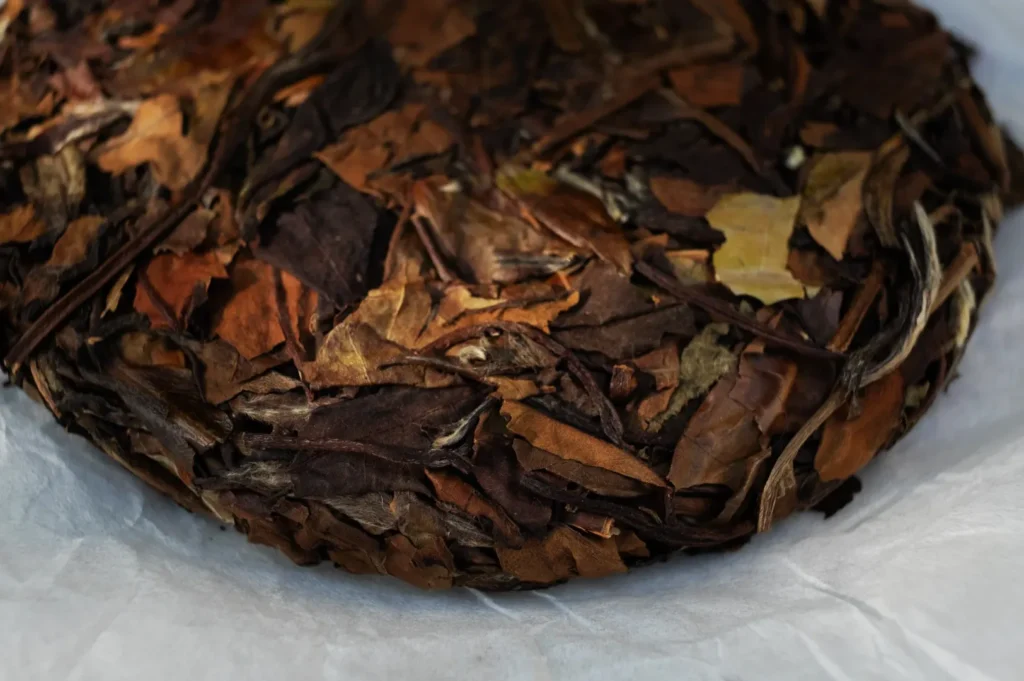 A round, compressed cake of dried tea leaves in shades of brown, placed on white paper. The texture is rustic, evoking a traditional and organic feel.