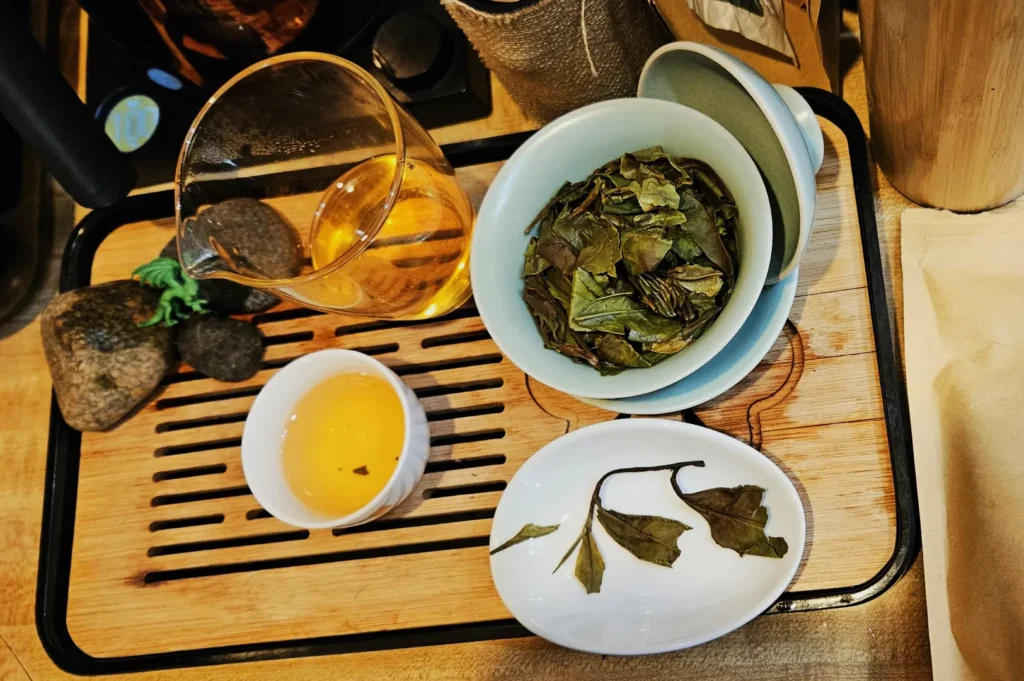 Wooden tray with a glass pitcher of golden tea, a bowl of loose green tea leaves, a cup of tea, and a plate with a few loose leaves, creating a calming tea setup.