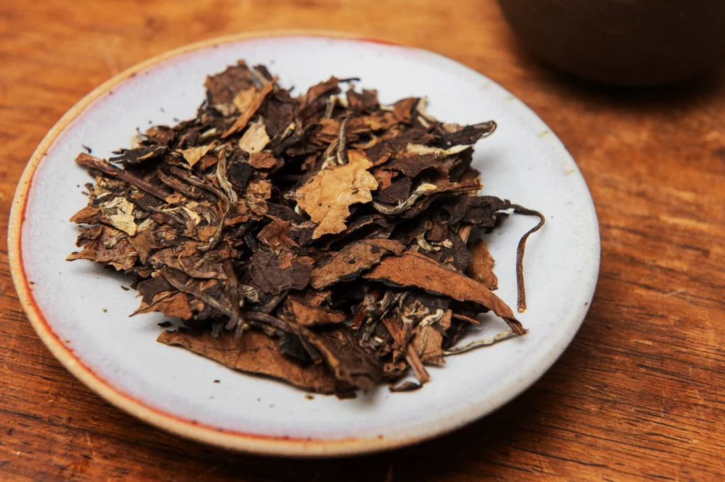 Dried tea leaves on a white ceramic plate with a reddish rim, resting on a wooden surface. The scene conveys a rustic and organic feel.