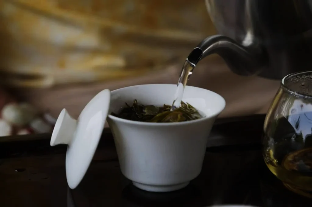 A teapot pours hot water into a white cup filled with tea leaves. The scene conveys a calm, ceremonial atmosphere with subdued lighting.