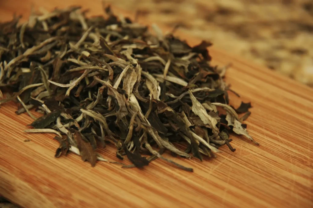 A pile of loose leaf white tea with silver-green leaves on a wooden cutting board, conveying a natural, earthy feel. Close-up shot, warm tones.