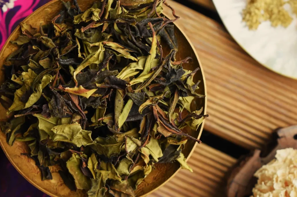 A close-up of dried tea leaves in a wooden bowl on a slatted surface. The leaves are varied shades of green and brown, suggesting a rustic, earthy tone.