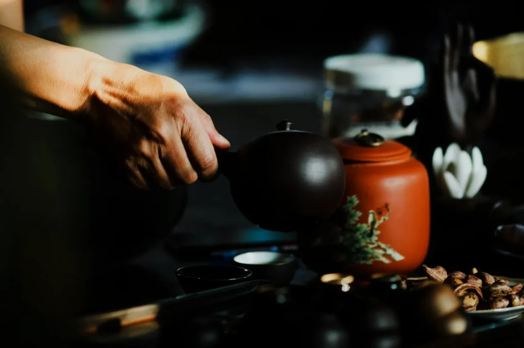 A hand pouring tea from a dark teapot into a cup. An orange teapot with floral designs sits nearby. The scene is calm and intimate with soft lighting.