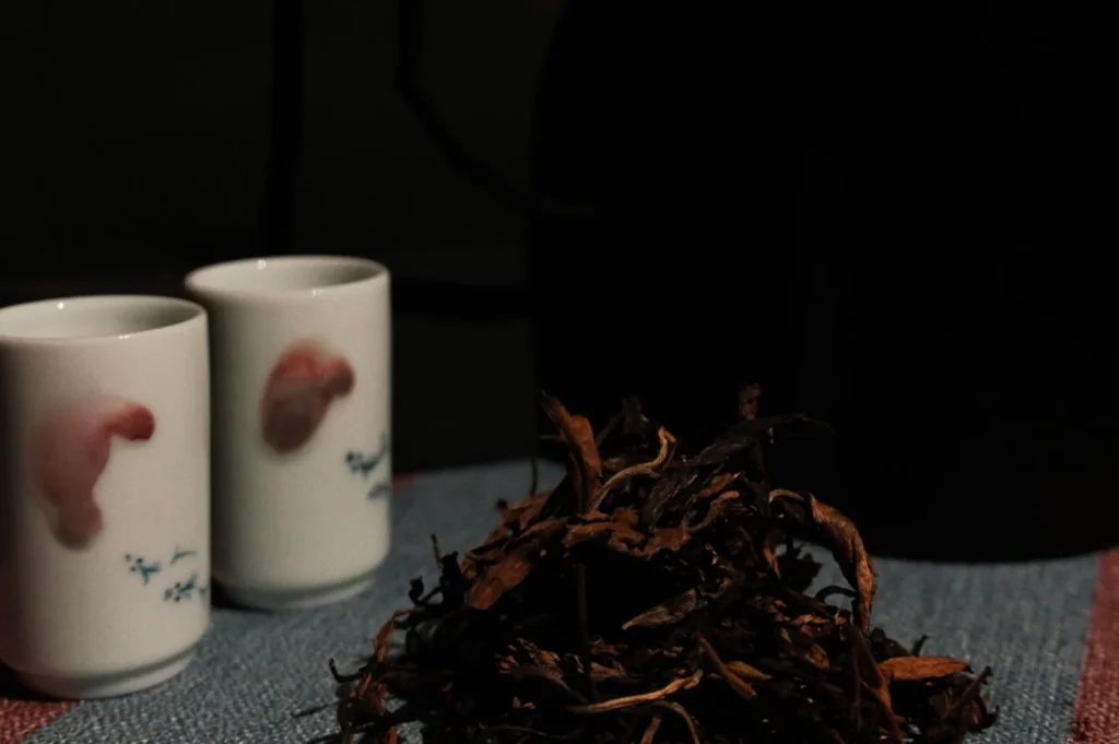 Dimly lit setting with two white ceramic cups featuring pink designs, placed on a cloth next to a pile of dried tea leaves, creating a cozy, serene mood.
