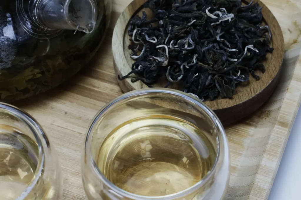 Two glass cups filled with light yellow tea on a wooden tray beside loose, dark tea leaves on a round wooden coaster, exuding a calm, natural vibe.