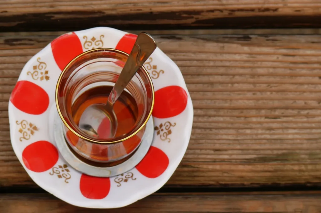 A glass of tea with a silver spoon rests on a red and white patterned saucer, placed on a wooden table. The scene conveys a warm, relaxed atmosphere.
