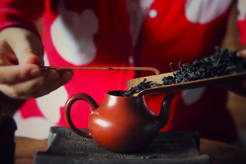 Hands carefully pour loose tea leaves from a wooden scoop into a small, brown teapot. The person wears a red sweater with large white spots.