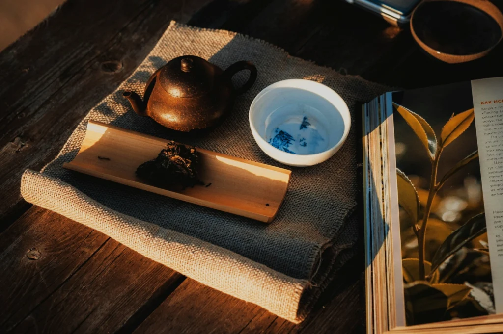 Rustic tea setup on a wooden table with a small teapot, white cup, wooden tray with tea leaves, burlap cloth, and an open book, evoking a peaceful, cozy vibe.