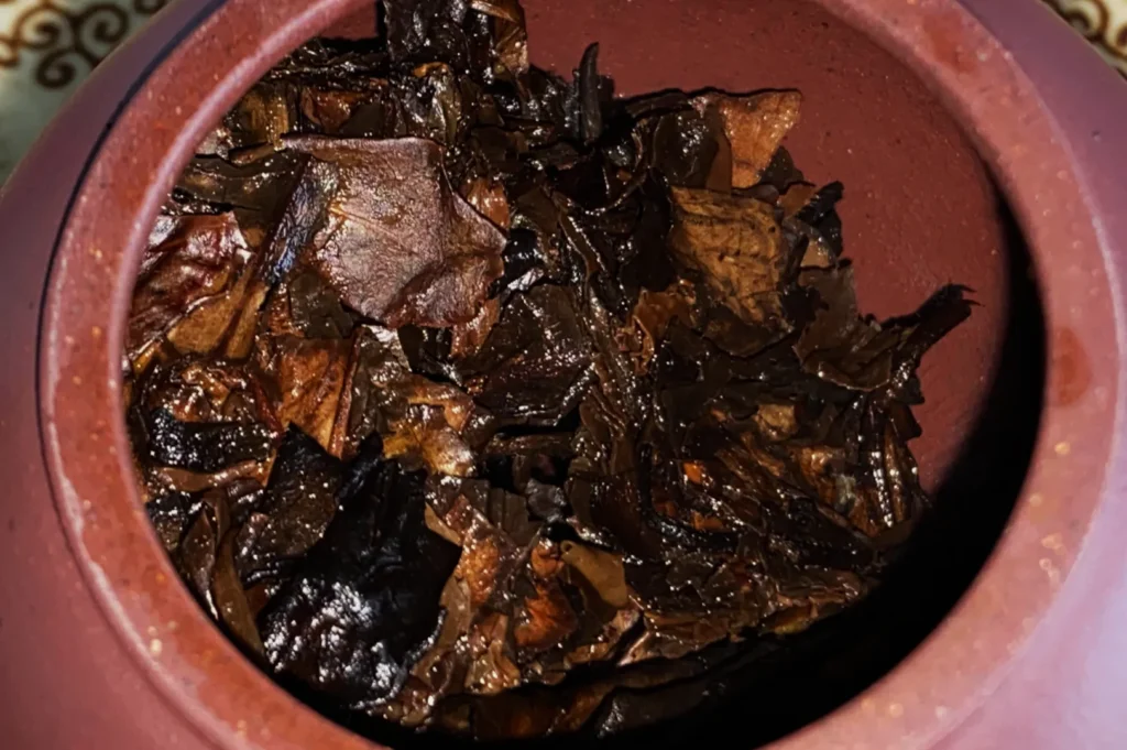 Close-up of wet, dark brown tea leaves inside a round, clay teapot. The leaves appear shiny and textured, suggesting freshness and richness.