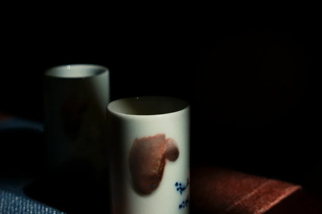 Two ceramic cups with subtle floral designs are partially illuminated in a dark setting. The mood is calm and contemplative, with soft lighting.