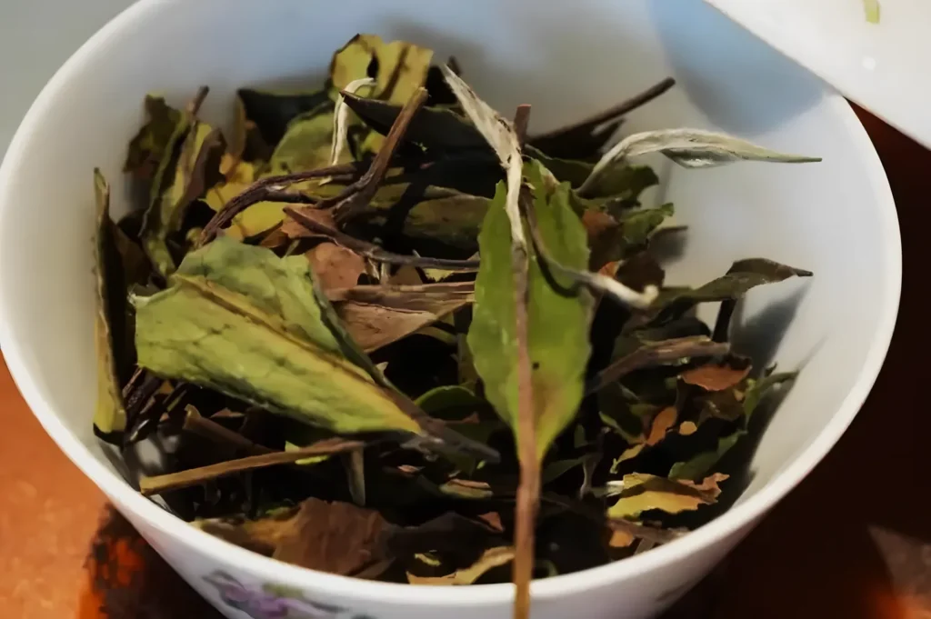 A close-up of dried tea leaves in a white teacup. The leaves are green and brown, varying in size and shape, conveying a natural, earthy tone.