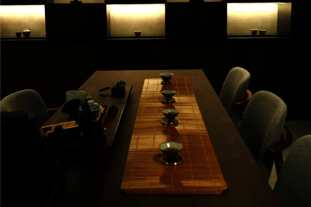 Dimly lit tea room with a long wooden table set for a tea ceremony. Five small cups are neatly arranged, creating a calm and serene ambiance.