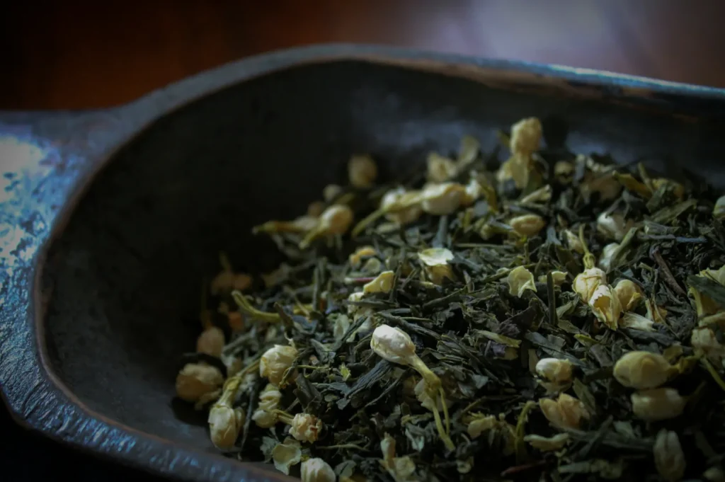 A rustic metal dish holds a blend of dried green tea leaves and jasmine flowers, creating an earthy and calming tone. The focus is close-up and detailed.