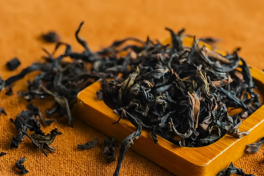 Loose-leaf black tea is scattered on a wooden tray against an orange fabric background. The scene conveys a rustic, earthy tone.