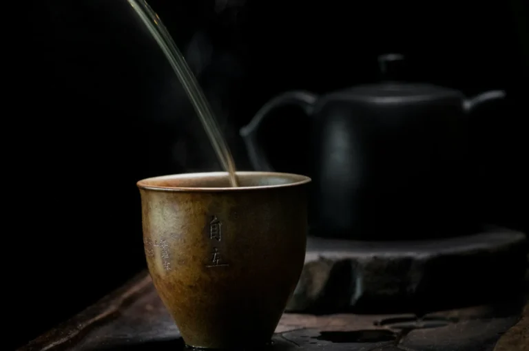 A stream of tea being poured into a rustic, brown cup with Asian characters, set against a blurred, dark teapot in the background, evoking calm.
