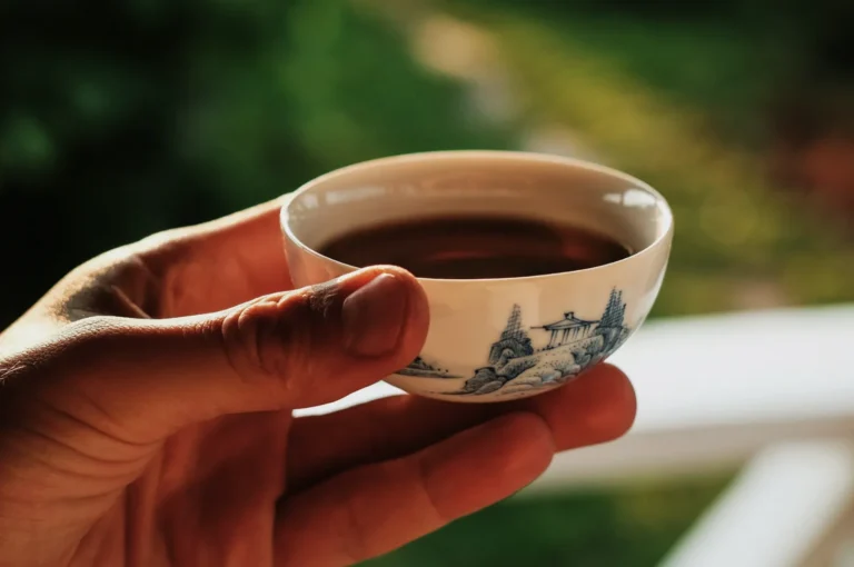 A hand holds a small, ornate teacup filled with tea against a blurred outdoor background. The cup features delicate, blue landscape designs.