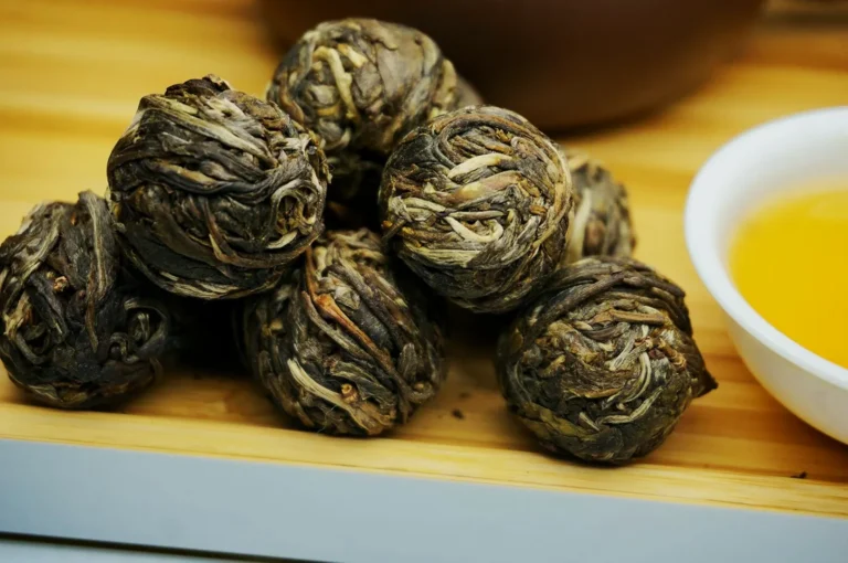 Tea balls made from dried tea leaves are neatly arranged on a wooden surface. A small white bowl filled with golden tea is beside them, creating a cozy feel.