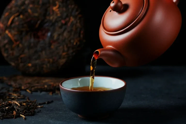 A brown teapot pours tea into a black and white cup on a dark surface, with loose tea leaves and a tea cake in the background, creating a calm, traditional atmosphere.