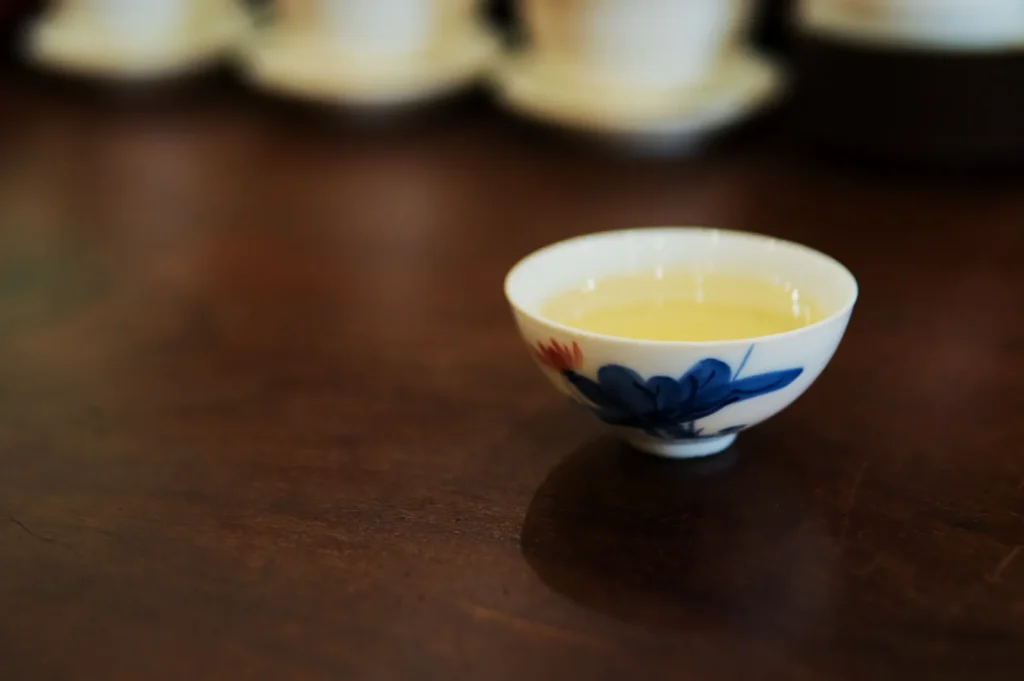 A small, elegant teacup with blue floral patterns, filled with light tea, sits on a wooden table. The background is softly blurred, creating a warm ambiance.