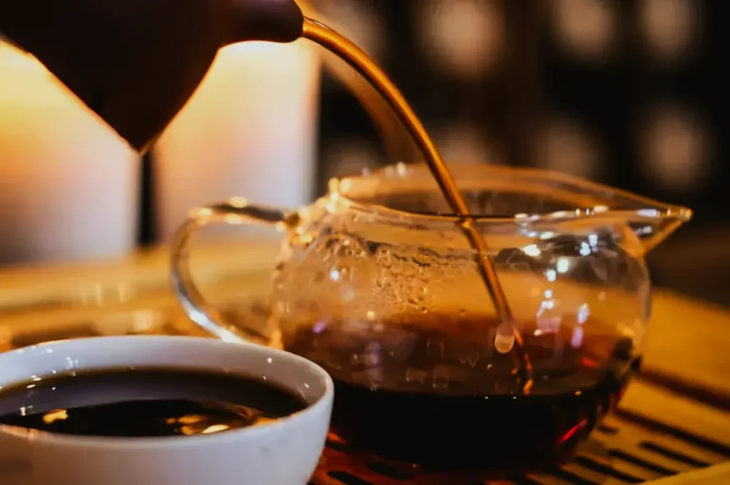 A person pours dark tea from a clear teapot into a white cup. The setting is warm and cozy, with soft candlelight in the background.