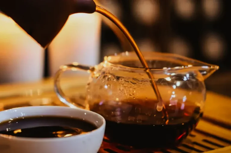 A person pours dark tea from a clear teapot into a white cup. The setting is warm and cozy, with soft candlelight in the background.