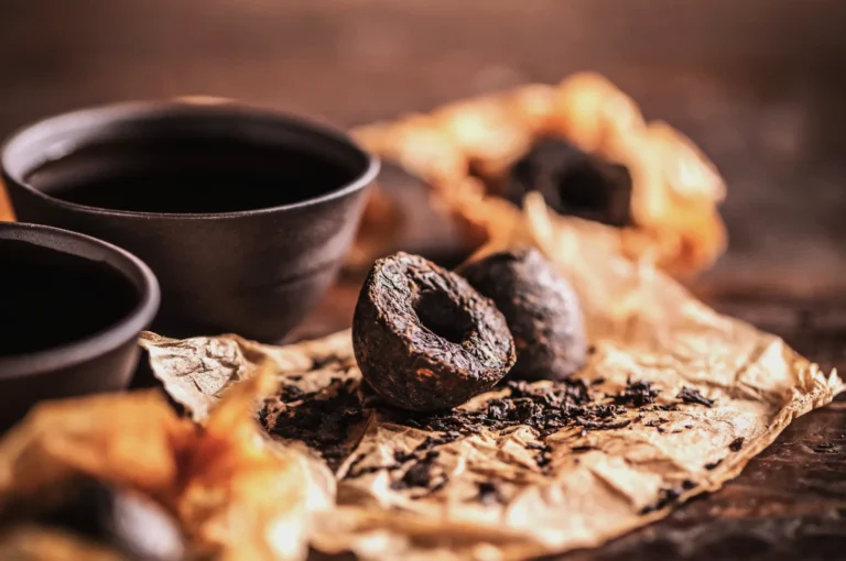 Two cups of tea next to dried Pu-erh tea cakes on crumpled paper. The rustic setting and warm tones convey an inviting and cozy atmosphere.
