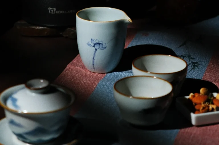 A serene scene features a floral-painted tea pitcher and cups on a textured cloth. Soft shadows and gentle lighting create a calming ambiance.