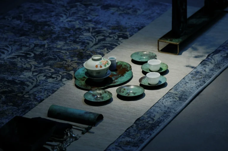 Elegant tea set arranged on a textured blue and beige fabric. Features a white teapot, small cups, and rustic greenish-blue plates, creating a serene atmosphere.