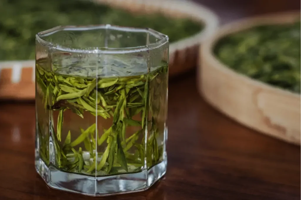 A clear glass filled with fresh green tea leaves in water, set on a wooden table. In the background, more tea leaves are drying on trays, creating a calm, organic atmosphere.